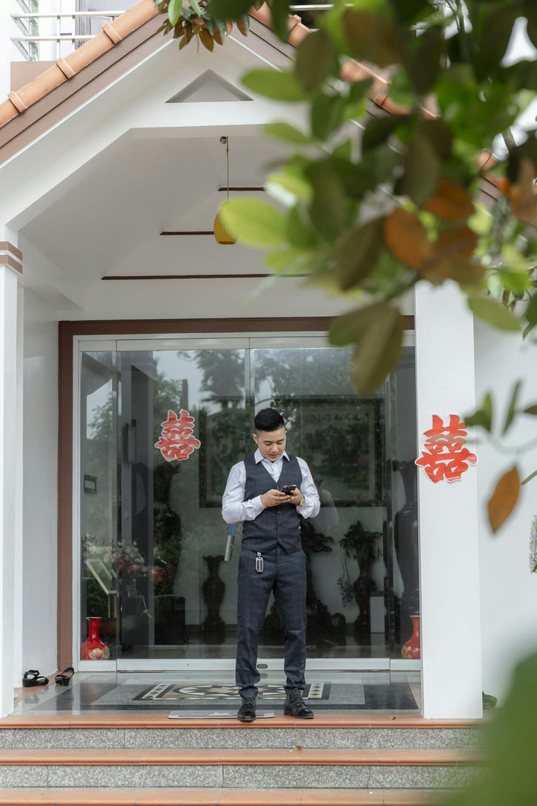 A groom stands stylishly outside a modern house, ready for the big day, dressed in a formal suit.