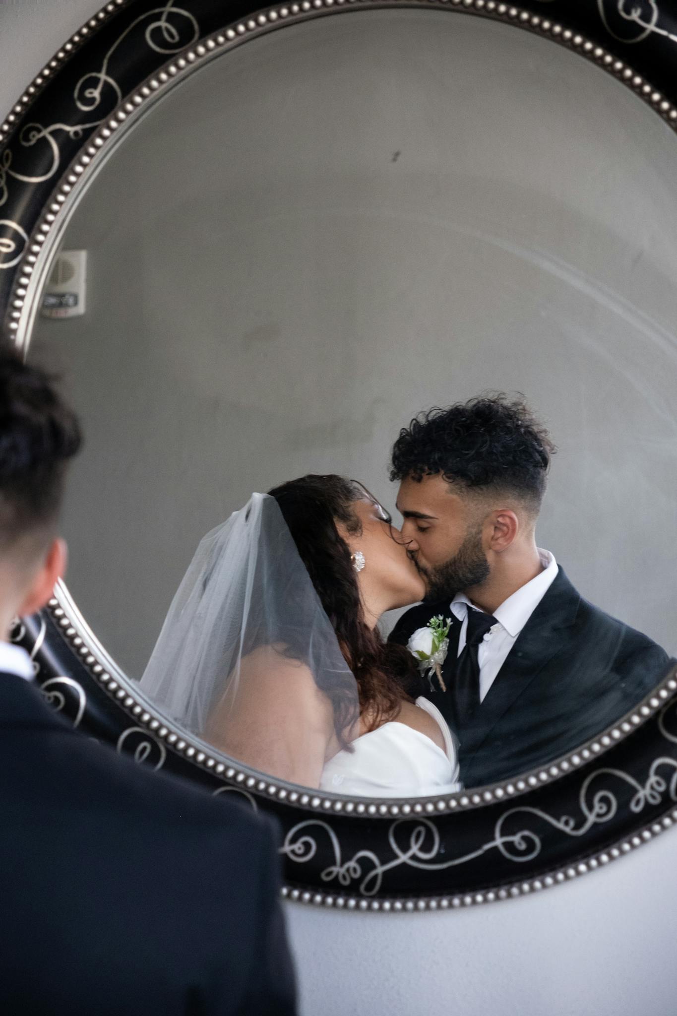 A newlywed couple shares a romantic kiss captured through a decorative round mirror, symbolizing love and togetherness.